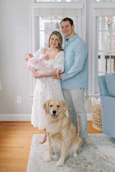 Richmond Family Photographer | Photo of a mom and dad holding their newborn baby girl swaddled in a pink bow as their dog looks on by Jacqueline Aimee Portraits