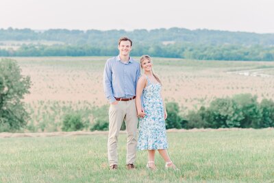 An engaged couple stands on a mountain top with a valley below holding hands in blue