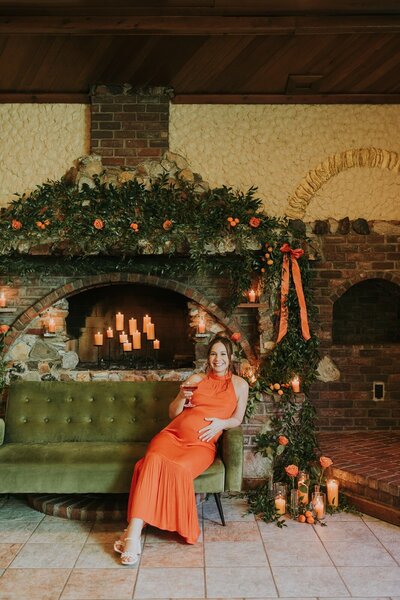 Pregnant woman in an orange dress sitting on a green couch in front of a decorated brick fireplace with greenery, orange roses, and candlelight, in a warm, rustic setting in century room at grbic events in st. louis