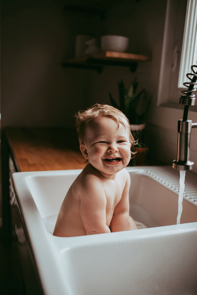 Toddler sitting in farmhouse kitchen sink bath with toothy smile.