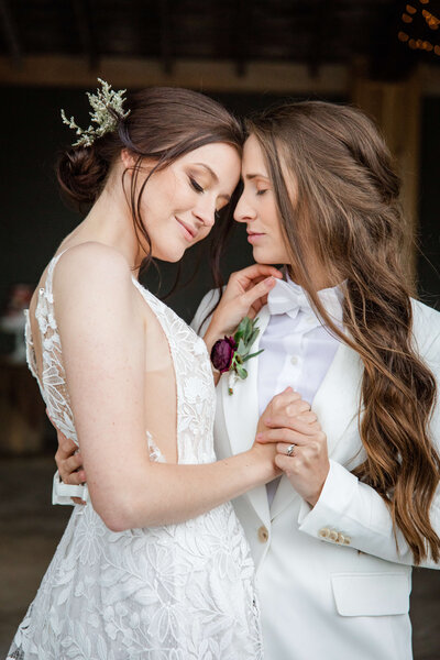 A woman and another woman hold hands and hug. One woman is in a wedding dress and the other is in a white suit. They are smiiing with their eyes closed.