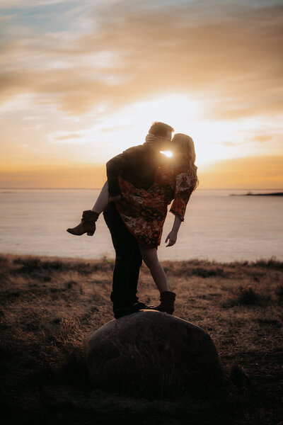 A man and his fiancée kiss while standing on a rock in a grassy field with the sun setting behind a lake behind them