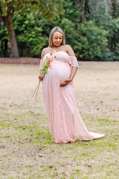 Mom to be holding flowers and looking down at her belly in a peach lace maternity gown in Lexington SC