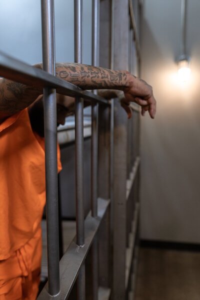 A man with tattoos on his arms wearing an orange jumpsuit inside a prison cell