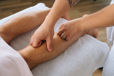 Close-up of a therapeutic leg massage using deep thumb pressure, performed by a therapist with a visible arm tattoo.
