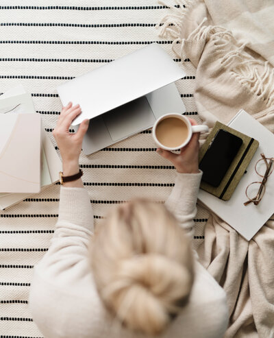 Frau sitzt mit Kaffee in der Hand auf dem Boden in der anderen Hand hat sie einen Laptop