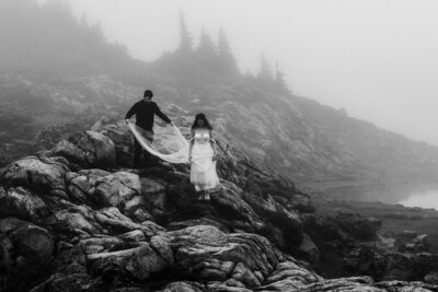 a black and white image of a couple exploring a rocky and foggy landscape in their wedding attire. 