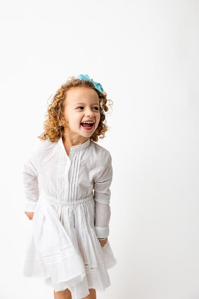 Girl in white laughs at camera in a white photo studio.