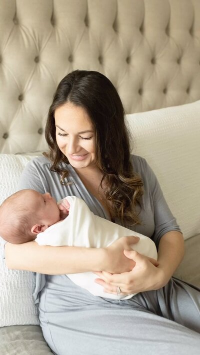 Mother in a soft gray dress gazes at her swaddled newborn with love. A warm in-home newborn session by Moments in Light Photography.