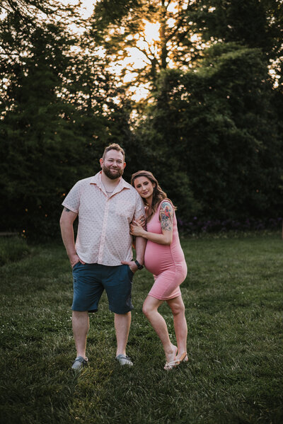 Maternity photo of Mom hugging dads arm while wearing pink and blue in front of trees and beautiful golden setting sun.