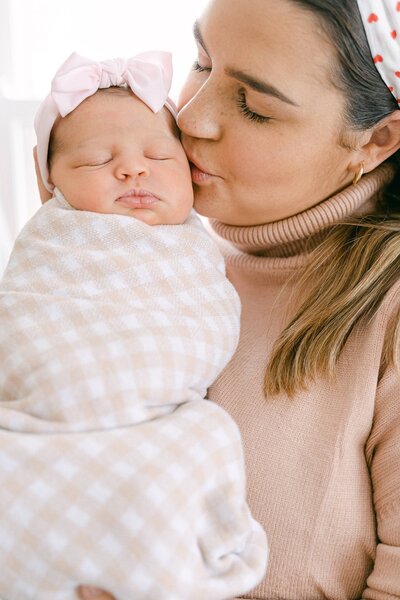 mother kissing her newborn baby girl wrapped in a beige gingham swaddle