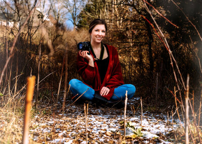 Girl is sitting cross legged with snow scattered on the ground in front of her and bushes and trees behind her. She's hold a camera in one hand and smiling at the camera.