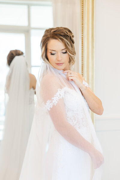Bride wears a San Antonio wedding dress and veil while looking over her shoulder down at the ground in front of a mirror. The dress includes an off the shoulder sleeve with lace.