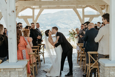 A bride and groom kiss each other after their wedding ceremony. Joyous guests celebrate around them.