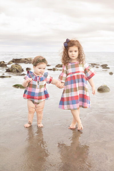 beach portrait of sisters wearing matching plaid dresses; the sky is overcast and the sea is gray, punctuated by the rocks of the shoreline