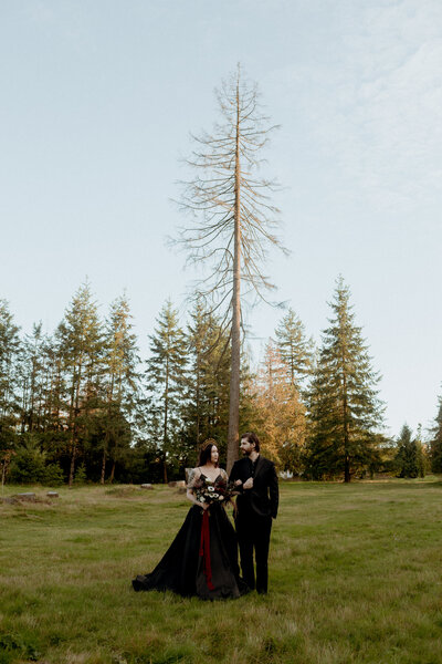 Couple in their black wedding outfits looking at each other with a tall tree behind