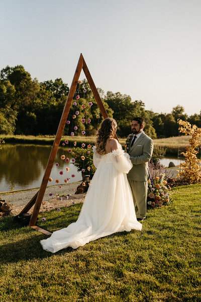 Outdoor wedding photography captures a romantic moment with a bride in an elegant white gown and a groom in a tailored olive suit. They stand before a triangular floral arch adorned with vibrant flowers, set against a serene lake backdrop. The bride's flowing dress with a long train and the groom's stylish suit add a touch of elegance to the natural setting, complete with lush greenery and golden sunset lighting.