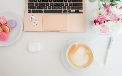 overview of white table with laptop, airpods, coffee, flowers and macaroons