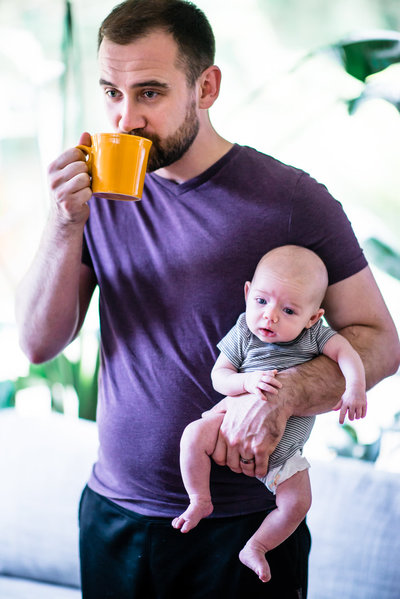 Dad drinking coffee in yellow mug while holding newborn