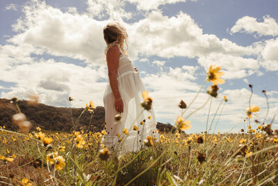 A person in a white dress stands in a field of yellow flowers under a partly cloudy sky, evoking the serene beauty of Umina Beach.