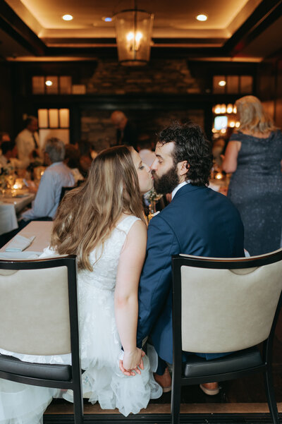 A newlywed couple shares a kiss while seated at their wedding reception in an intimate, warmly lit venue. The bride, wearing a lace wedding gown, has long, wavy blonde hair cascading down her back, while the groom, dressed in a navy blue suit, leans in with his curly hair and beard. They hold hands behind their chairs, adding a tender, unspoken connection to the moment. The softly glowing lanterns above create a cozy atmosphere as guests in the background enjoy their meals and conversations. The romantic, candid nature of this moment encapsulates the joy and intimacy of their special day.