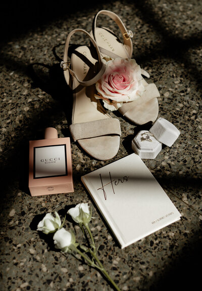 Close up of a brides wedding rings, florals, perfume, shoes, and vow book on the ground with a window shadow across it.
