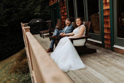 Bride and Groom joyfully holding hands and sitting in wooden Adirondack chairs at their mountain Airbnb after eloping.