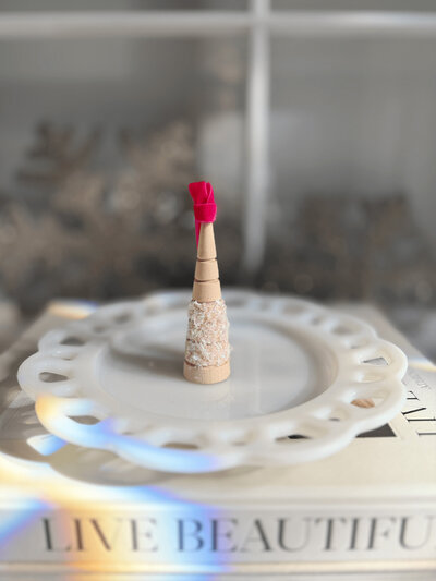 A small, cone-shaped holiday ornament decorated with beige crystals and topped with a pink spiral stands on a white decorative plate, which is placed on top of a book—a charming piece from Sweet Pea Ink Creative.