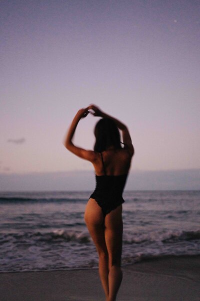 Meg O'Neill walking away on the beach with a black bathing suit on and hands in the air