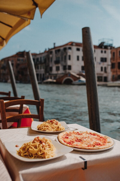 A table with pizza and pasta on the table riverside with buildings in the distance