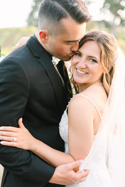 close-up image of groom kissing bride's cheek while she smiles at the camera in a sonoma vineyard.