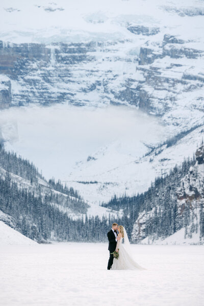 Bride and groom standing on the frozen surface of Lake Louise in winter with snow covered surroundings and Victoria Glacier in the background captured as a scenic wedding portrait