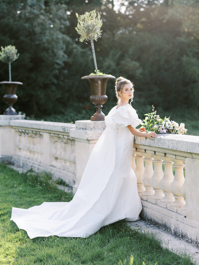 Bride in white gown with lace sleeves and veil holding blue and white bouquet against dark European mural backdrop at Cornman Farms