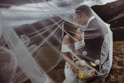 Bride and groom kiss under veil during cloudy Colorado mountain wedding.