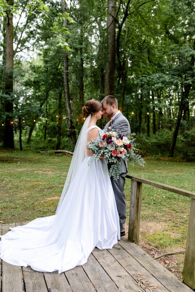 bride and her groom stand on a wooden bridge with their foreheads together with lighting strung in the woods behind them for their little rock ar wedding