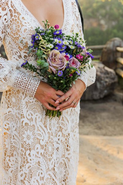 A bride is wearing a long sleeved crochet gown holding a bouquet of purple, pink and green wildflowers. 