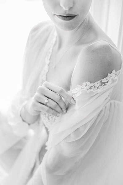 Richmond VA Boudoir Photographer | A black and white closeup image of a girl holding a sheer white robe off her shoulder. You can see her engagement ring as she holds the fabric.