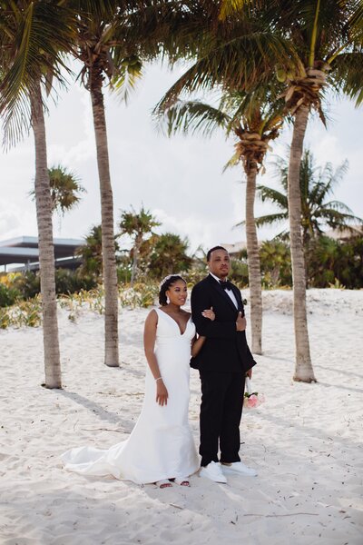 A bride and groom standing on the beach in front of palm trees.