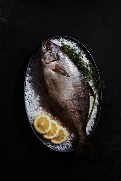 A striking image of a whole fish resting on a bed of ice, garnished with lemon slices and fresh herbs. Photographed by Mica McCook, this composition emphasizes the freshness and quality of seafood, presented in a minimalist style.