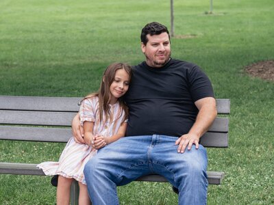 candid portrait of a father and daughter sitting on a park bench together. dad looks off to the side and the young girl leans in close to him in a tender moment.