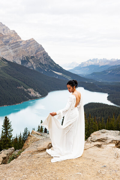 Beautiful bride staning on a mountain in Banff National Park. She is looking down on a blue lake in her wedding dress.
