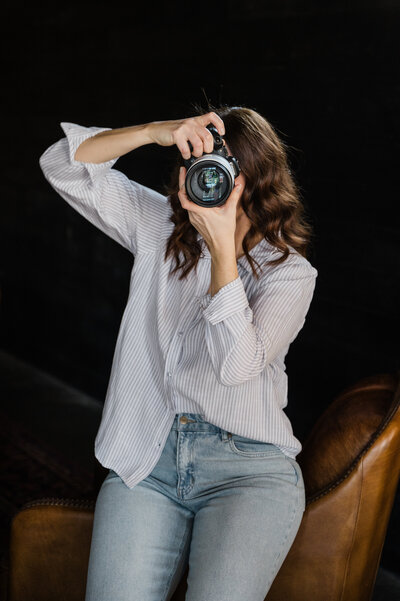 Woman holding a camera to take a picture perched on the arm of a leather chair wearing blue jeans and a light blue button up shirt