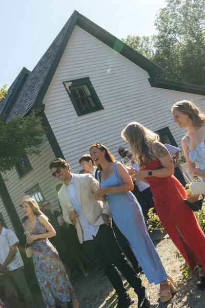 colourfully dressed wedding guests laugh in the sun beside farmhouse