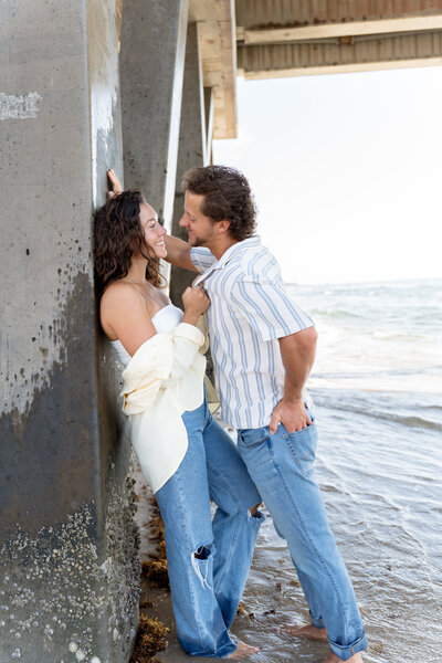 a couple on the beach is leaning against the pier. Looking at each other as she pulls him into her. Photographer is capturing the authentic and candid moment between them.
