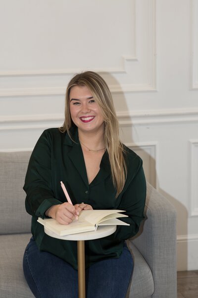 Karleigh Skeans sitting on a light gray couch, looking forward and smiling with a notebook and pen. She is wearing a dark green shirt and jeans.