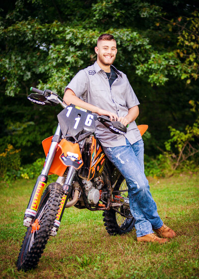High school senior boy standing against his dirt bike wearing jeans & a button-down, posing for a South Shore MA Senior photographer