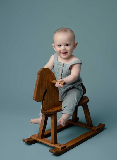 Baby boy in blue overalls, sitting on wooden rocking horse and smiling for 6 month milestone photo. Milestone baby photo taken at Atlanta Maternity photo studio in Woodstock GA.