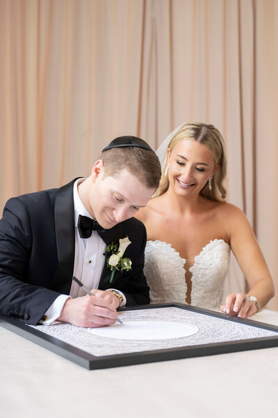 A bride and groom sit together at a table. The groom, wearing a black suit and kippah, signs a document, while the bride, in a strapless white dress, smiles by his side. A neutral-toned curtain hangs in the background.