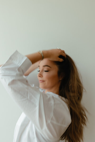 Alyssa Amez a custom wedding stationery and signage designer and business woman from Alyssa Amez Design posing in a white long sleeve button down shirt grabbing her hair from the side and smiling with her eyes closed