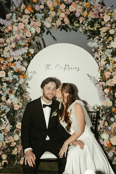 A bride kisses the groom’s cheek in front of a stunning floral arch, surrounded by the wedding party.